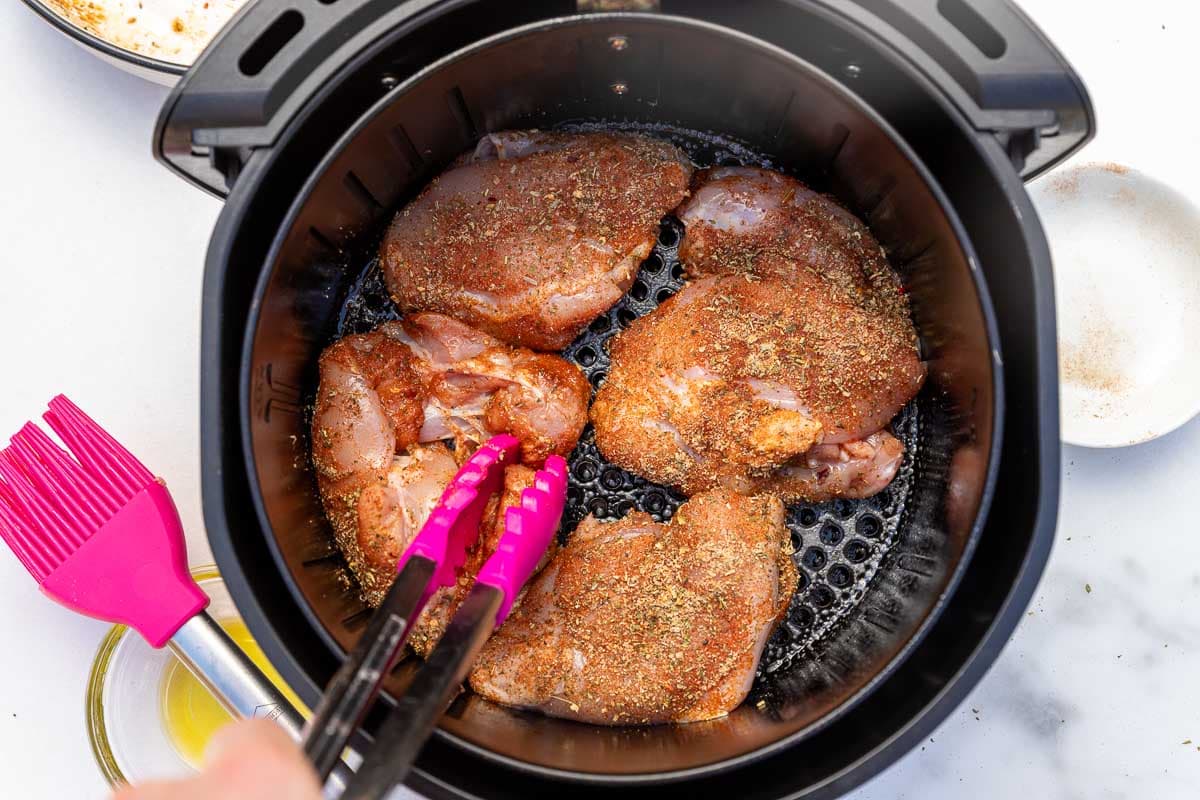 Overhead view of someone putting raw seasoned chicken thighs in an air fryer basket with pink silicon tongs.