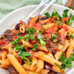 A closeup of the top of a dish of penne puttanesca pasta with fresh parsley on top and a spoon and fork in it.