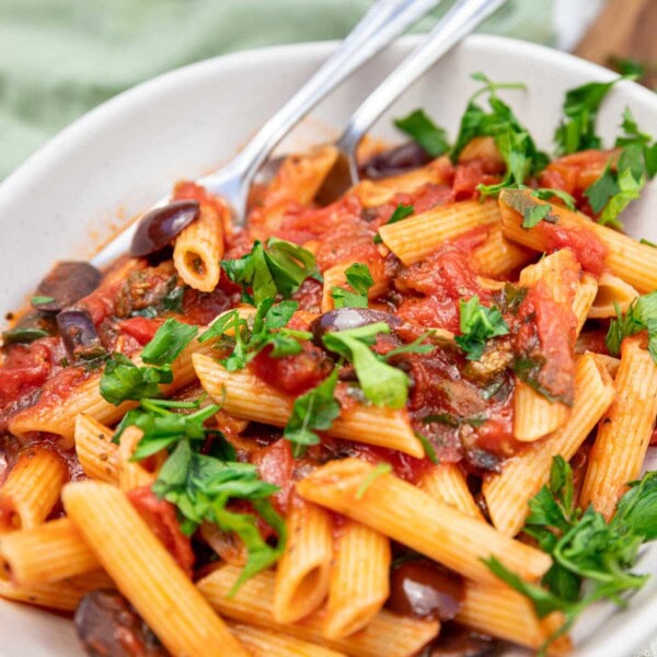 A closeup of the top of a dish of penne puttanesca pasta with fresh parsley on top and a spoon and fork in it.