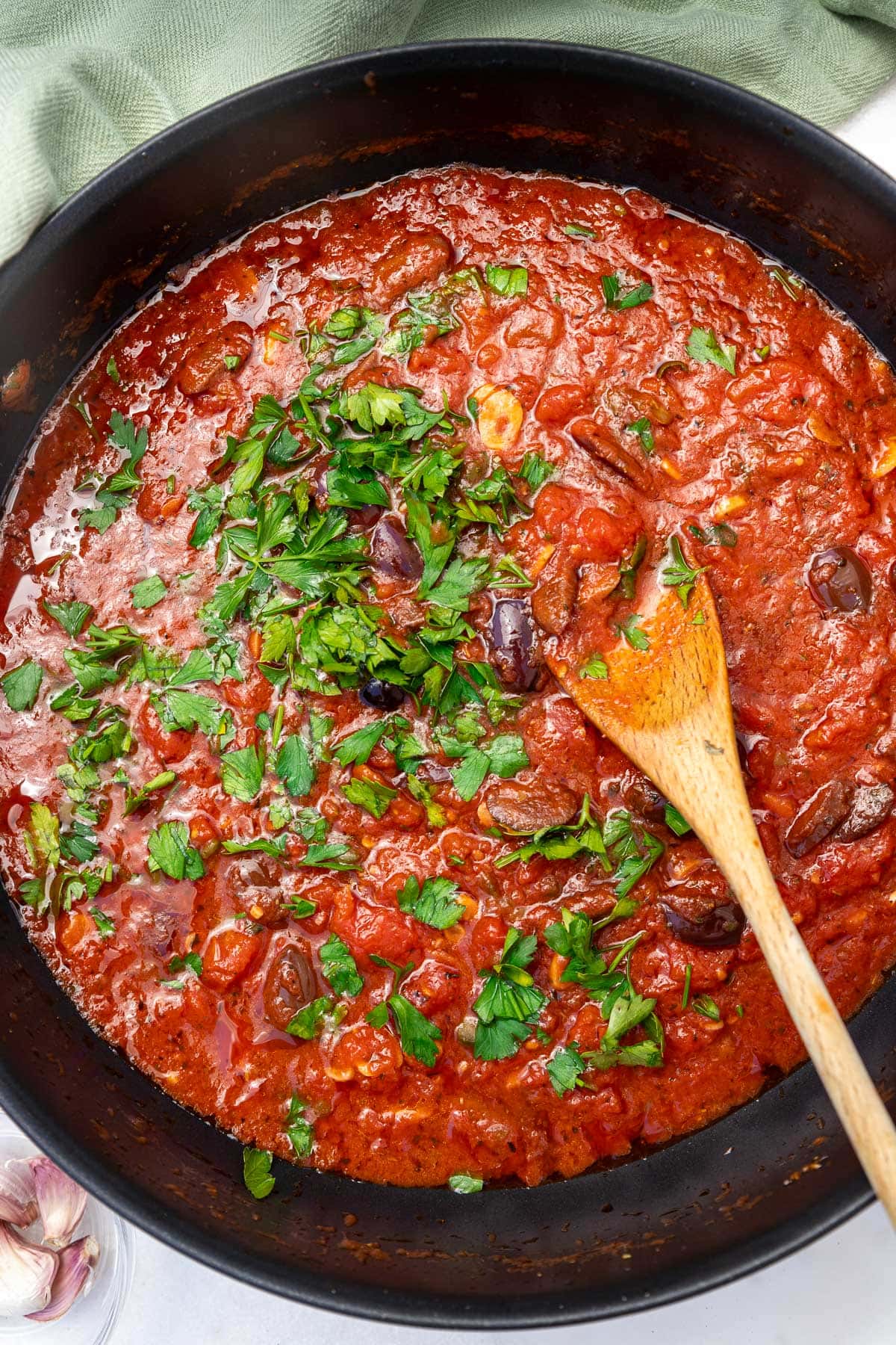 An overhead closeup image of a puttanesca pasta sauce in a large pan with parsley on top and a wooden spoon.