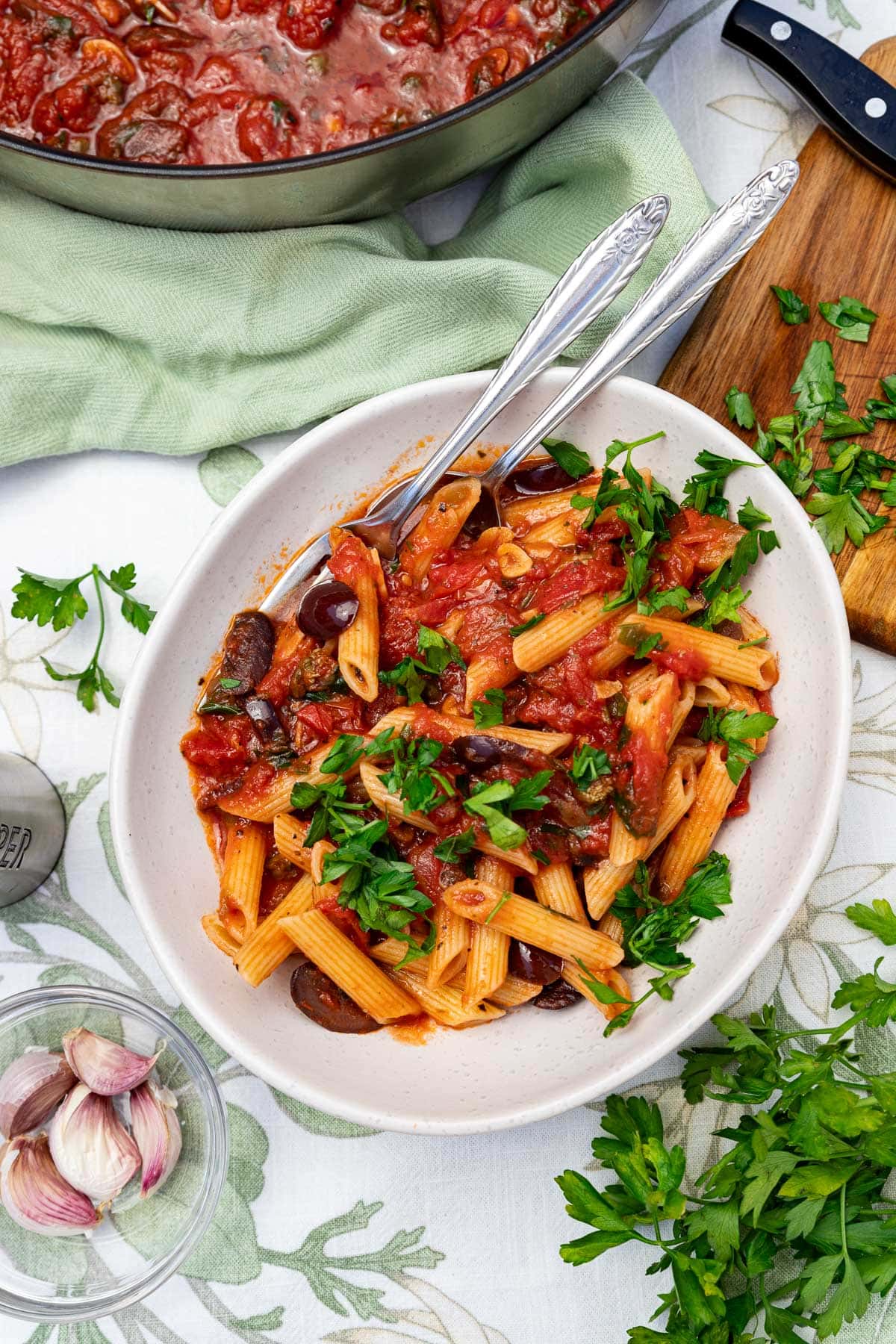 A serving of the pasta with fresh parsley on top on a green and white table cloth with a pan and ingredients around it.