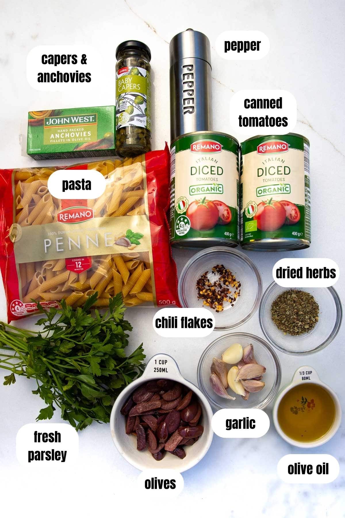 A labelled collage of all the ingredients you'll need including penne pasta, fresh parsley, canned tomatoes, olives, garlic, olive oil, dried herbs, chili flakes, pepper and anchovies and capers.