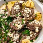 An overhead view of a white round dish of baked lamb chops with a simple Greek style marinade, with fresh herbs and feta on top and a green and white tea towel at the side.