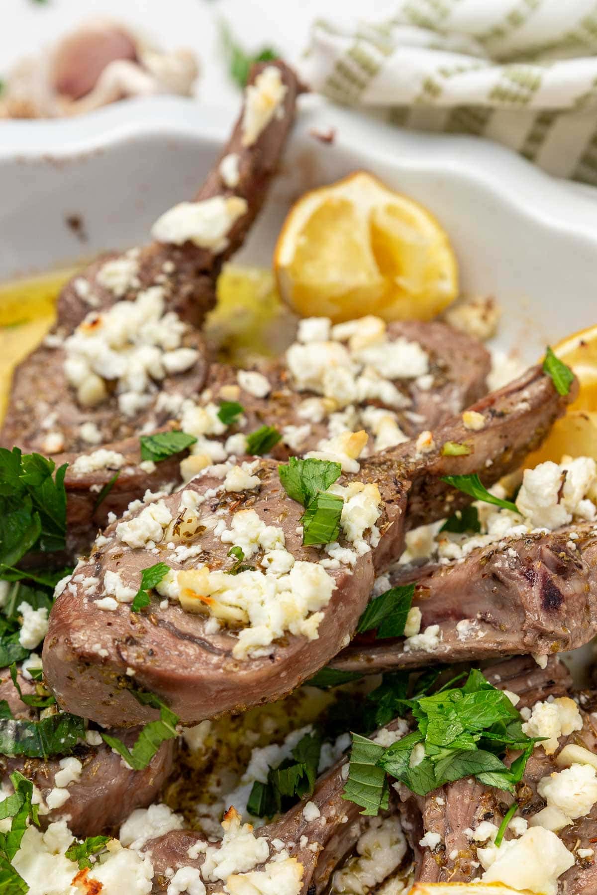 A closeup image of a Greek style oven baked lamb chop on top of many more, with fresh green herbs, feta and lemon on top.