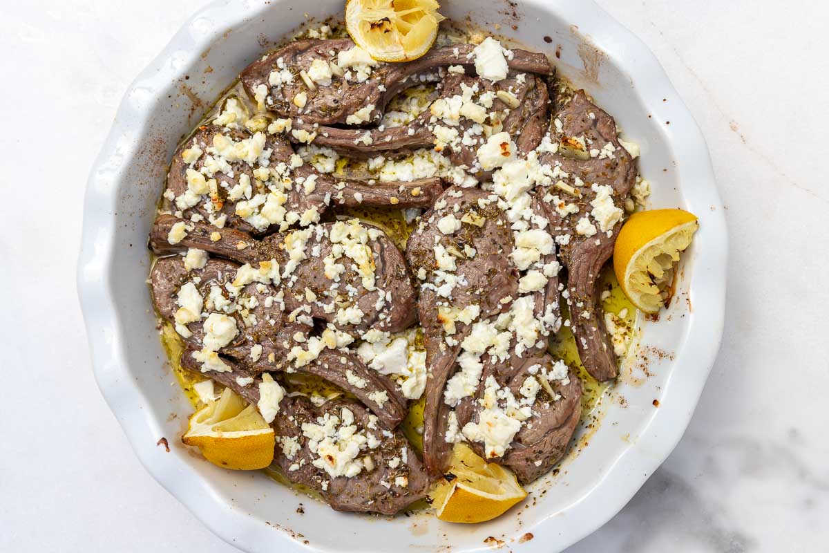 A round white baking dish with oven baked lamb chops with marinade, feta and lemon segments on top.
