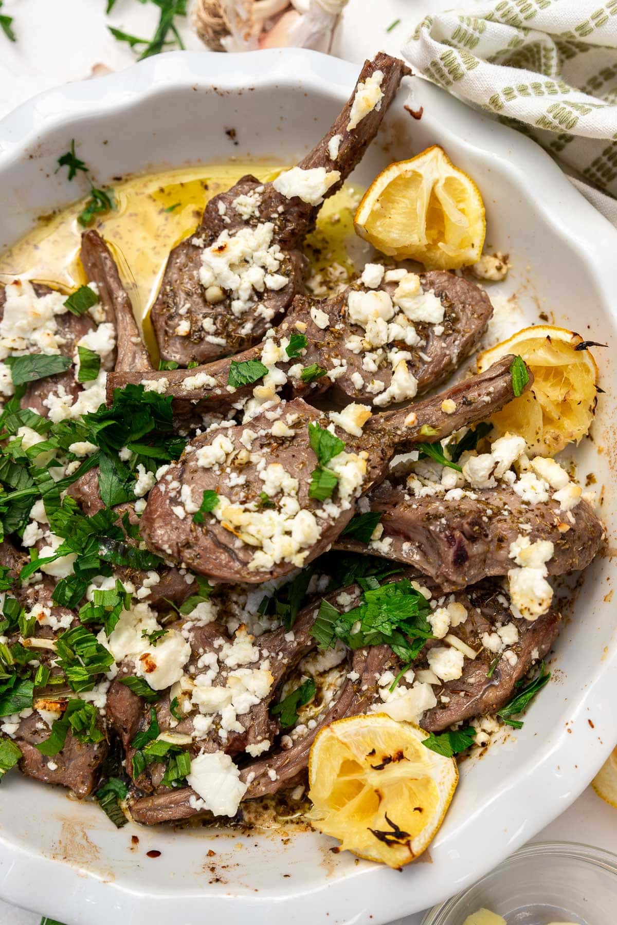 An overhead view of a white round dish of baked lamb chops with a simple Greek style marinade, with fresh herbs and feta on top and a green and white tea towel at the side.
