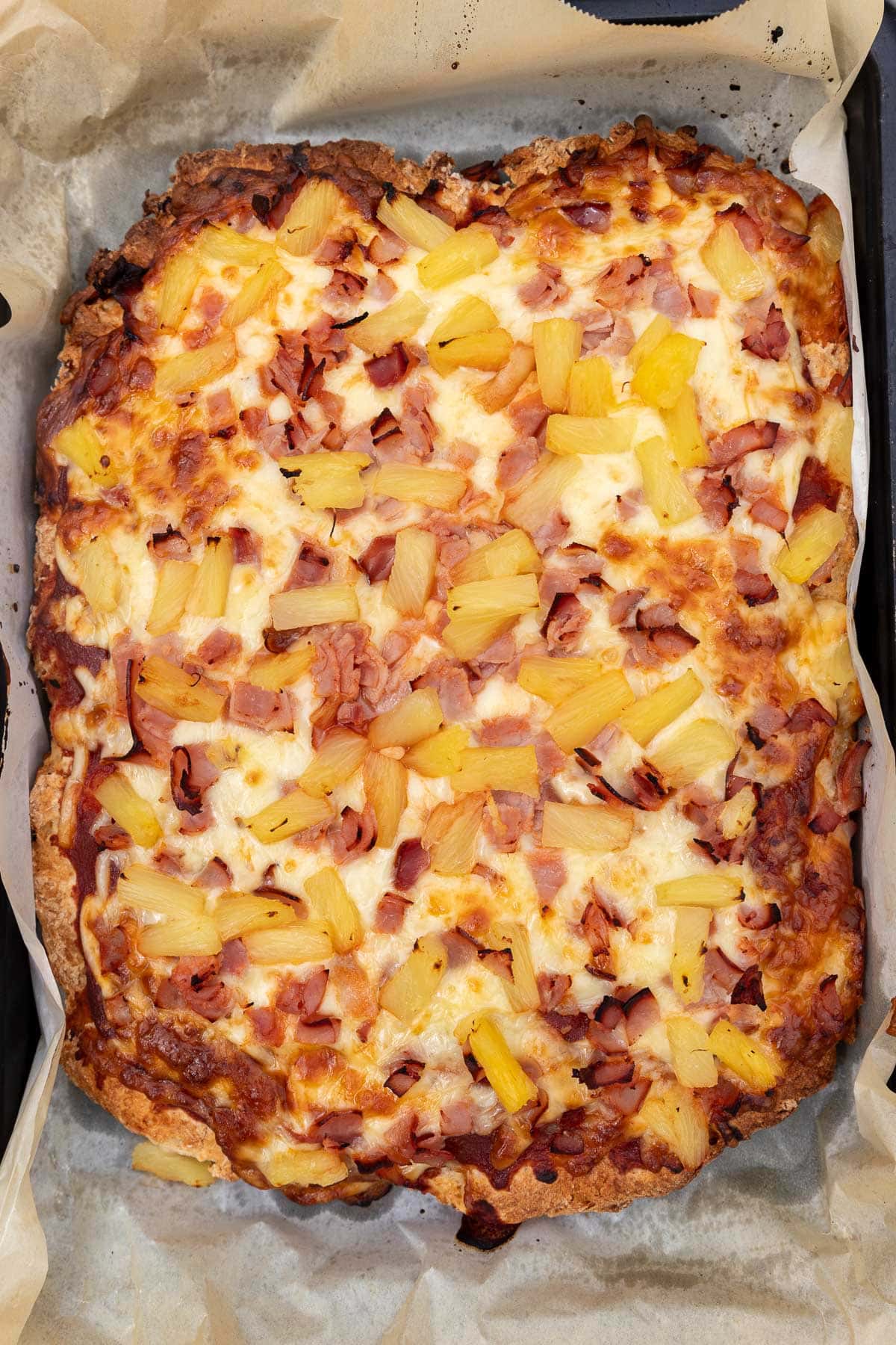 Overhead of just-cooked Greek yogurt ham and pineapple pizza on a baking paper lined baking sheet.
