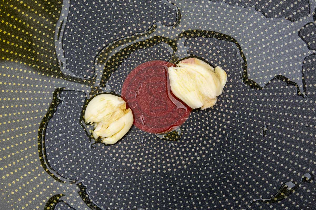 A closeup overhead of 2 squashed cloves of garlic in a pan with plenty of olive oil.