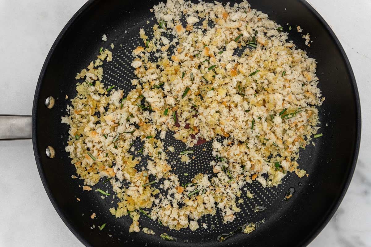 An overhead of breadcrumbs and rosemary mixed with olive oil in a frying pan, ready to be cooked and browned.