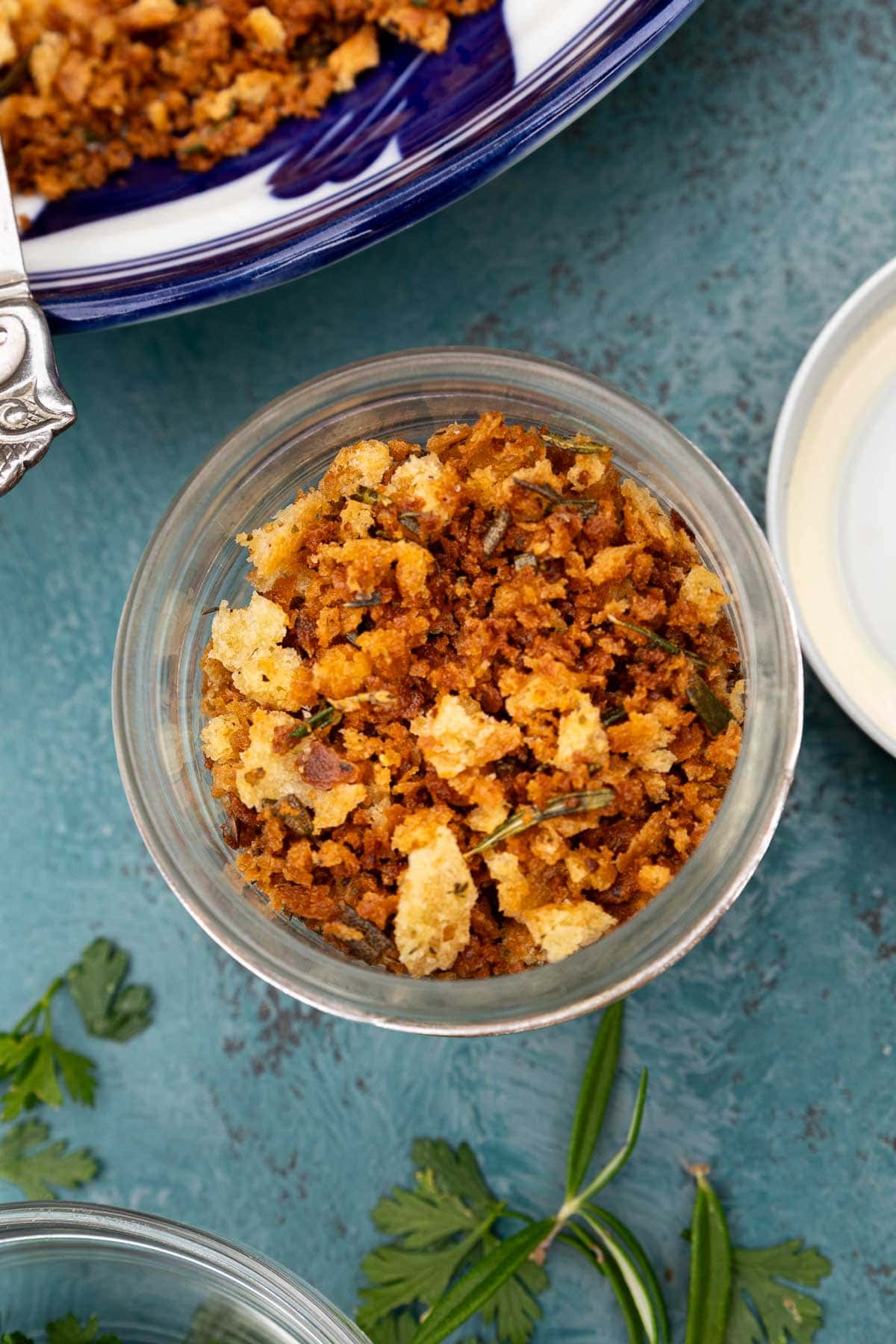 Overhead image of pangrattato or toasted Italian breadcrumbs in a small glass jar with the edge of the lid and herbs visible at the side.