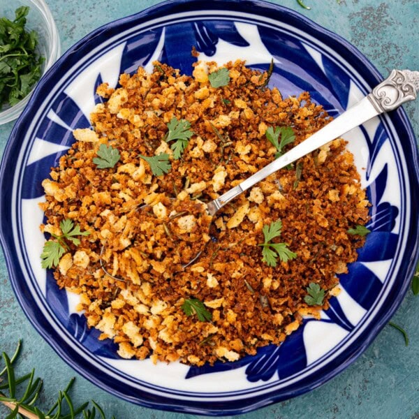 Italian toasted breadcrumbs, or pangrattato, in a patterned blue and white bowl with a decorative spoon on a blue background and with a sprig of fresh rosemary at the side.