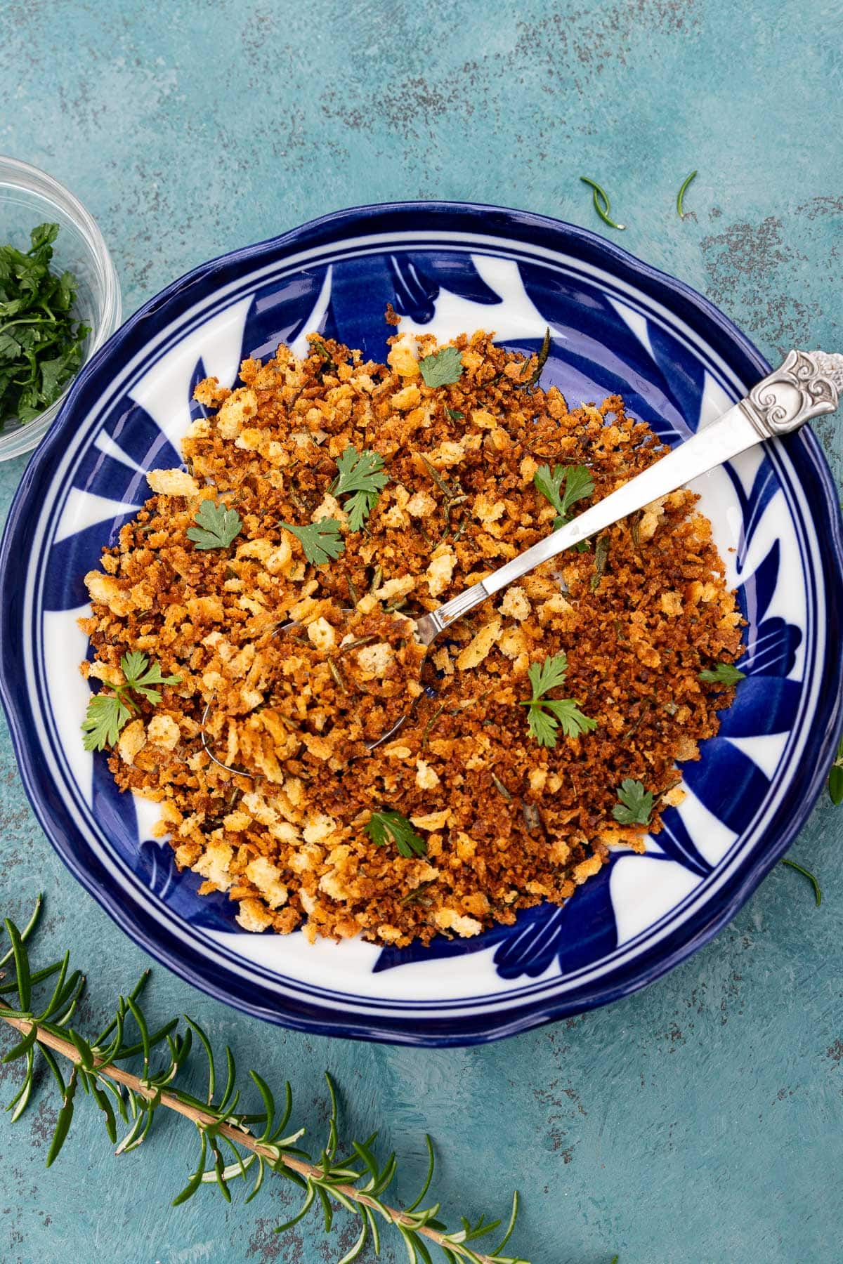 Italian toasted breadcrumbs, or pangrattato, in a patterned blue and white bowl with a decorative spoon on a blue background and with a sprig of fresh rosemary at the side.