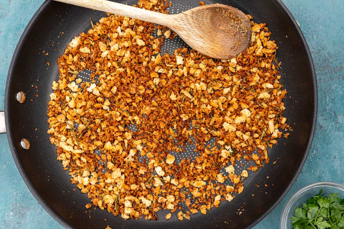 An overhead image of crispy Italian style breadcrumbs, or pangrattato, in a frying pan with a wooden spoon and on a blue background.