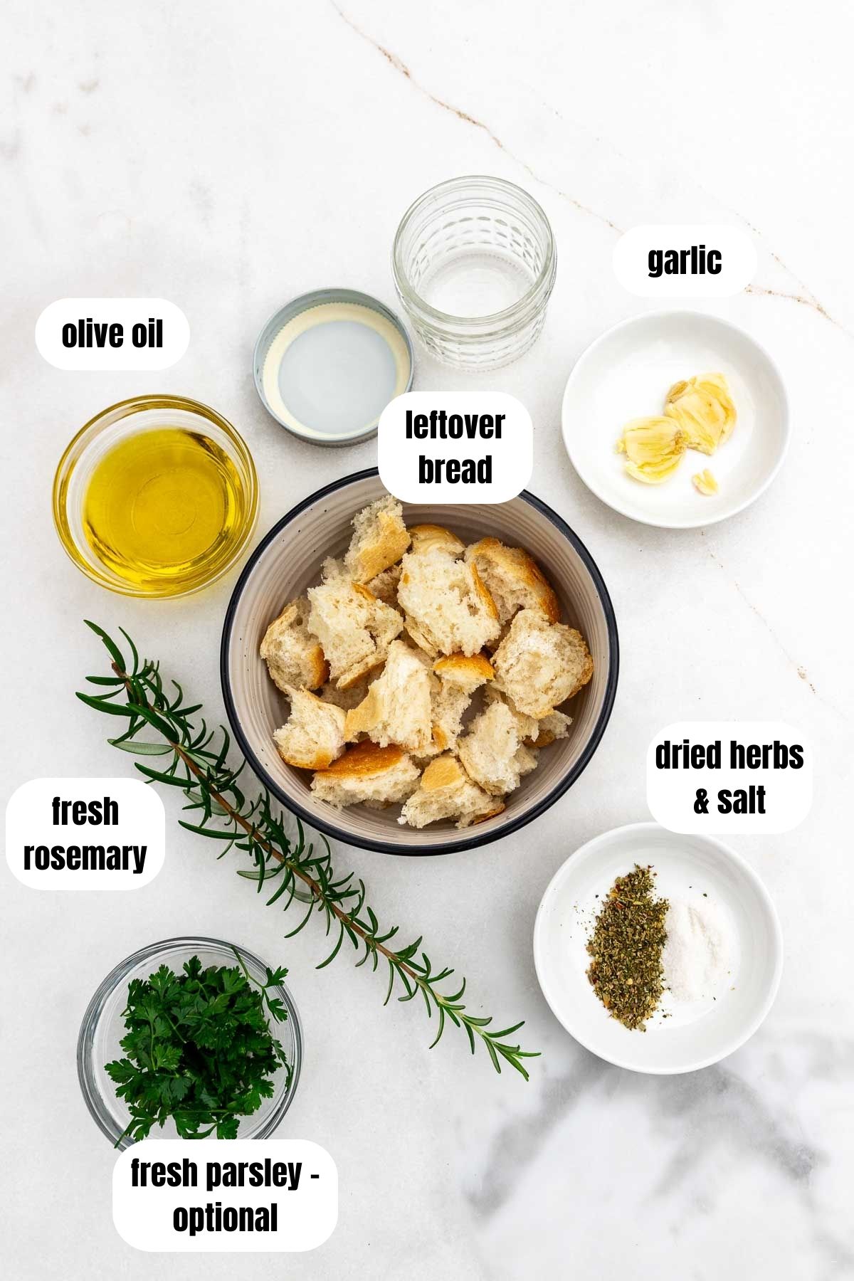 An overhead image of all the ingredients to make Italian crispy breadcrumbs, or pangrattato, labelled, including hunks of old bread, olive oil, dried herbs and salt, fresh rosemary and parsley, and two squashed cloves of garlic.
