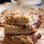 A closeup of a stack of 3 rhubarb bars on top of each other on a wooden board, with the rest in the background.