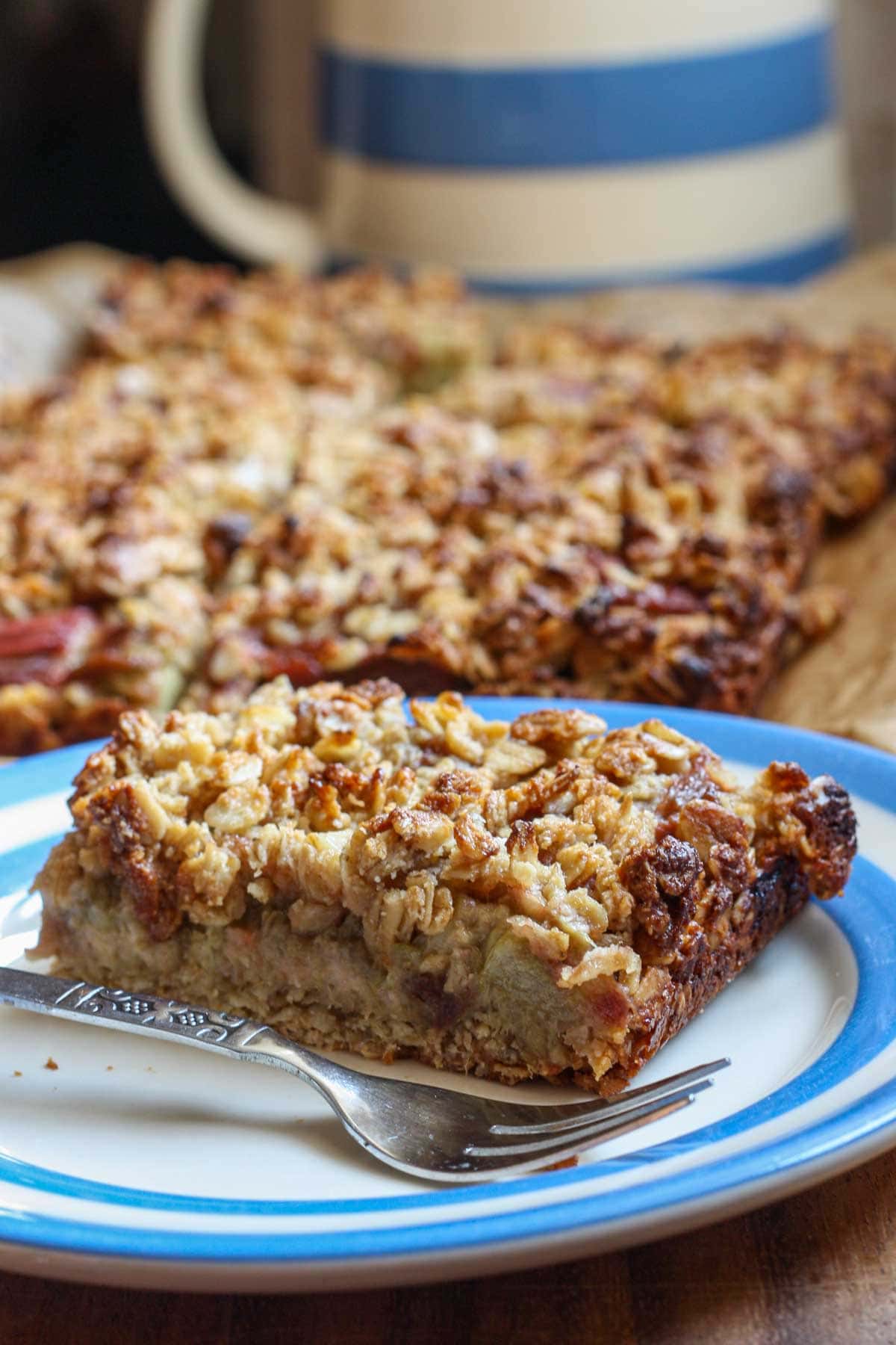 A rhubarb bar on a blue-rimmed plate with a fork, with the rest of the pan of bars in the background.