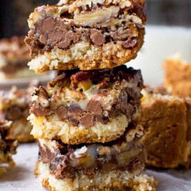A stack of three easy-to-make 7 layer bars with layers of graham cracker, coconut, chocolate, nuts and condensed milk on baking paper.