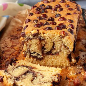 A Nutella loaf cake sliced so you can see the swirled inside, with whole hazelnuts on top and Nutella and milk in the background.