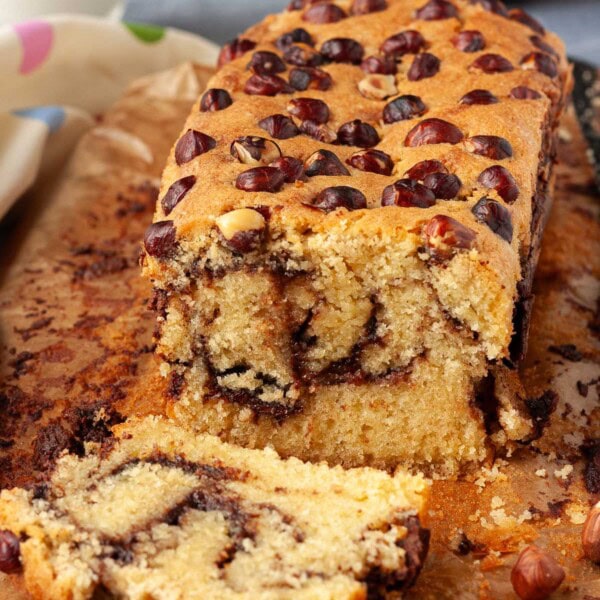 A Nutella loaf cake sliced so you can see the swirled inside, with whole hazelnuts on top and Nutella and milk in the background.