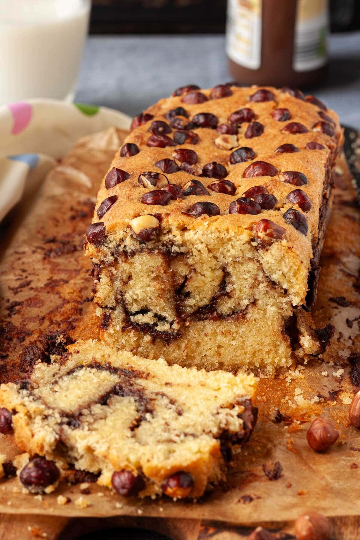 An easy Nutella cake sliced so you can see the swirls inside, with whole hazelnuts on top and Nutella and milk in the background.