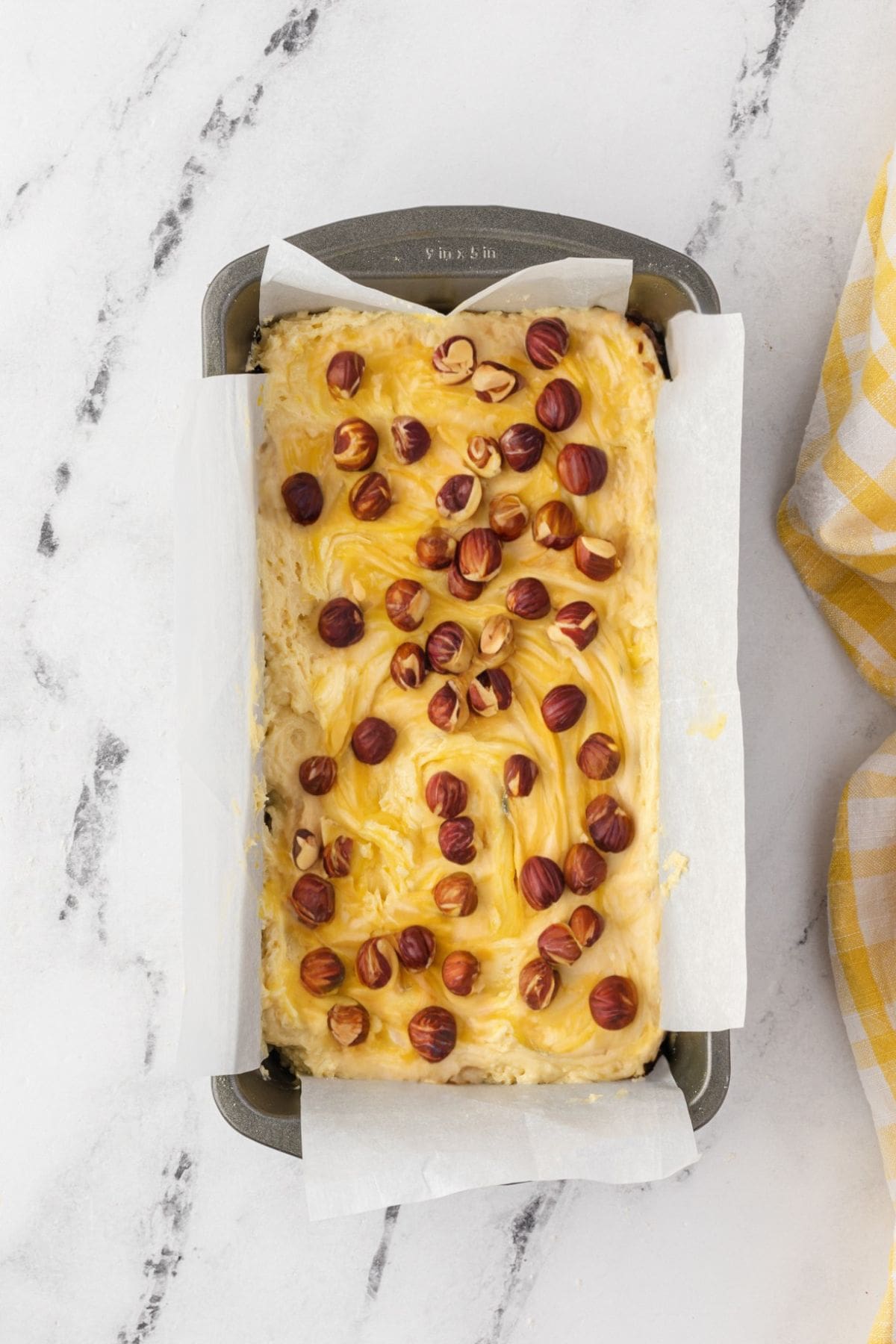 Raw cake batter in a loaf pan with whole hazelnuts on top.