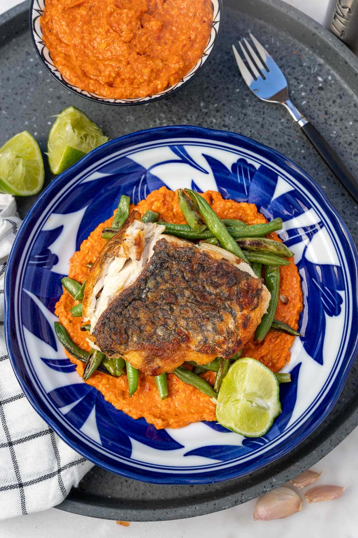 A blue and white patterned dish with romesco sauce, green beans, white fish and lime in it, with a checked tea towel and more sauce in a dish at the top.