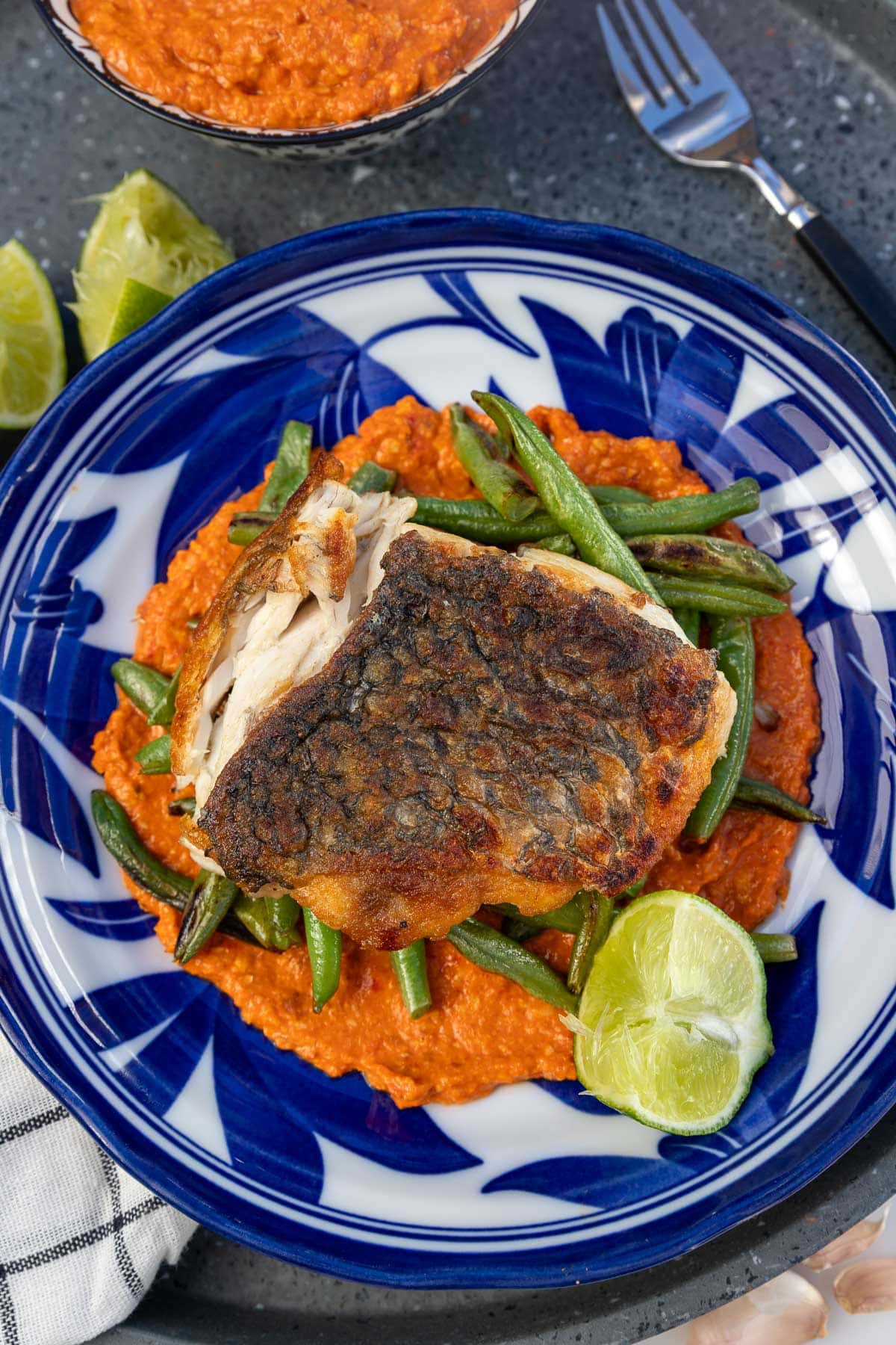 Closeup of a blue and white patterned dish with romesco sauce, green beans, white fish and lime in it, with a checked tea towel and more sauce in a dish at the top.