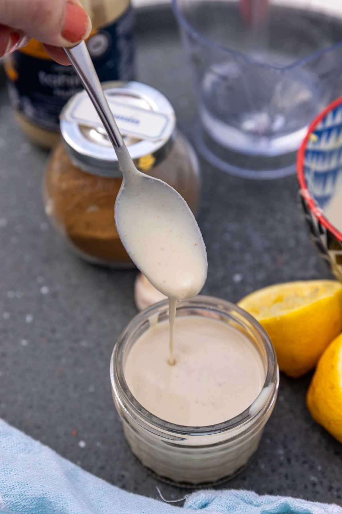 A teaspoon drizzling the dressing into a small jar, on a grey background and with ingredients next to it.