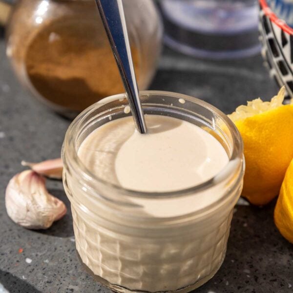 A small glass jar of lemon tahini salad dressing with a spoon in it and ingredients behind.