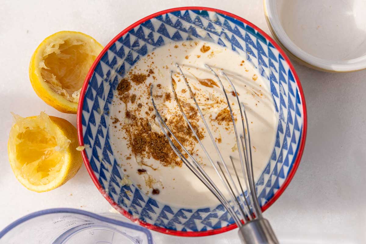 A small whisk in a small blue and red bowl of tahini and lemon juice, with a bit of cumin scattered over ready to be whisked in.