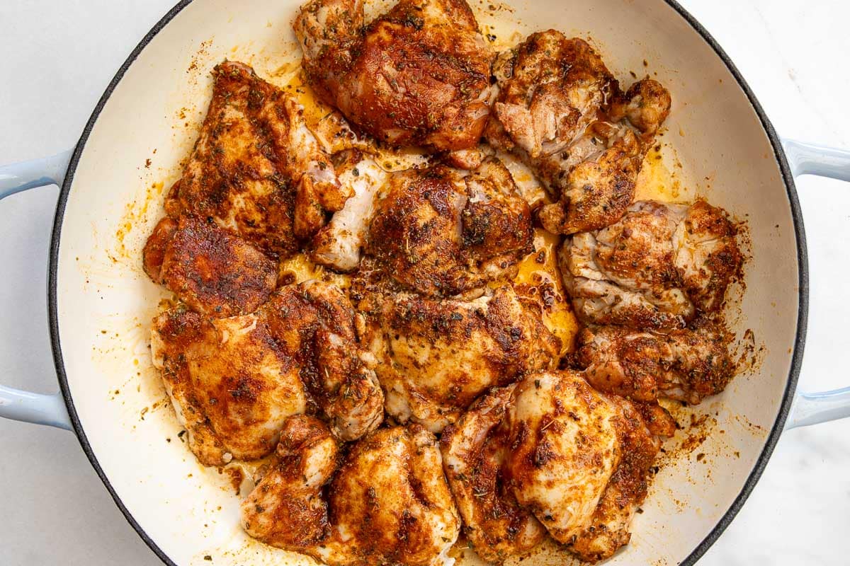 Overhead view of seasoned Greek chicken thighs covered in seasoning and browned in a wide cast iron pan.