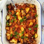Overhead view of a white with blue rim baking pan with roast potatoes tossed with tomato sauce and garnished with parsley, with a green tea towel and ingredients visible at the side.