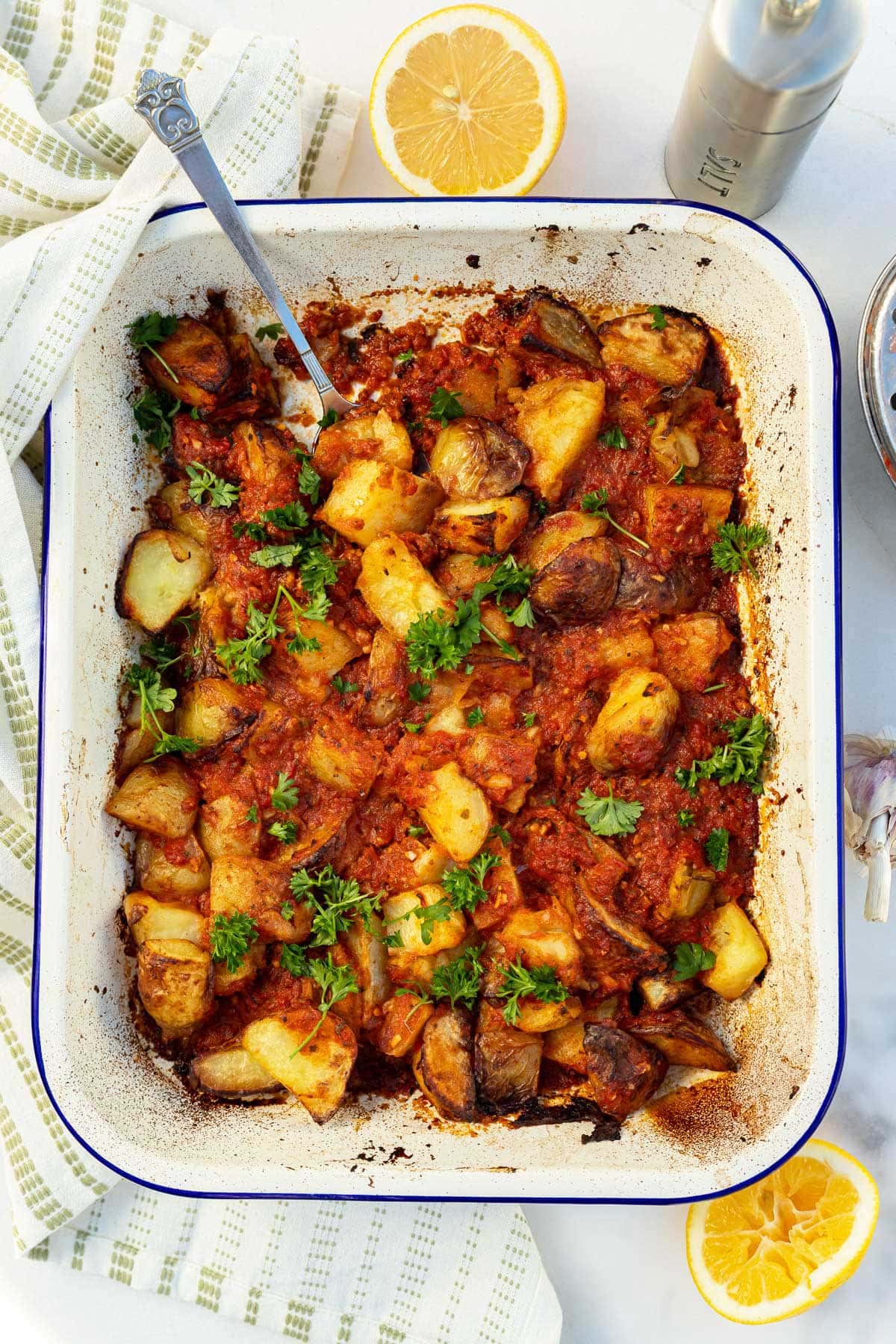 Overhead view of a white with blue rim baking pan with roast potatoes tossed with tomato sauce and garnished with parsley, with a green tea towel and ingredients visible at the side.