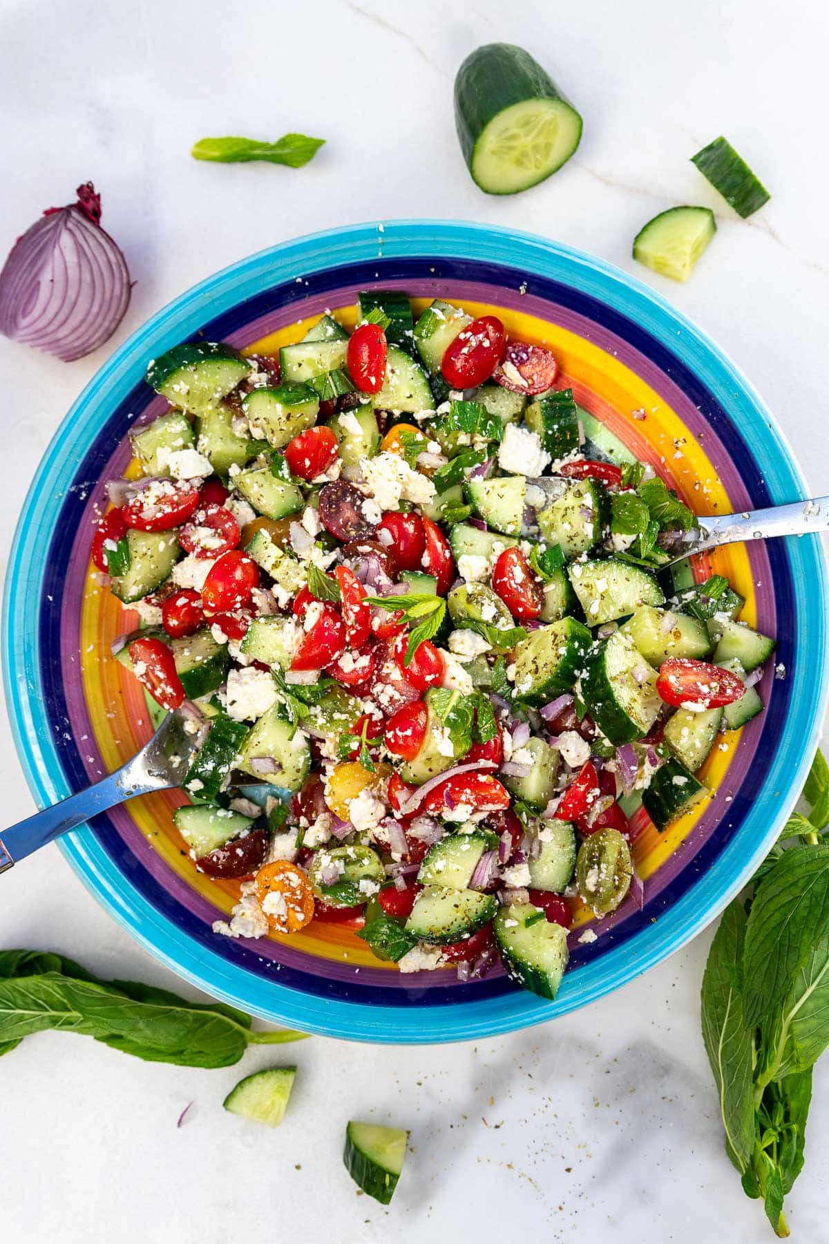 Overhead view of a rainbow colored bowl with the salad in it and salad servers, with some leftover ingredients around the side.