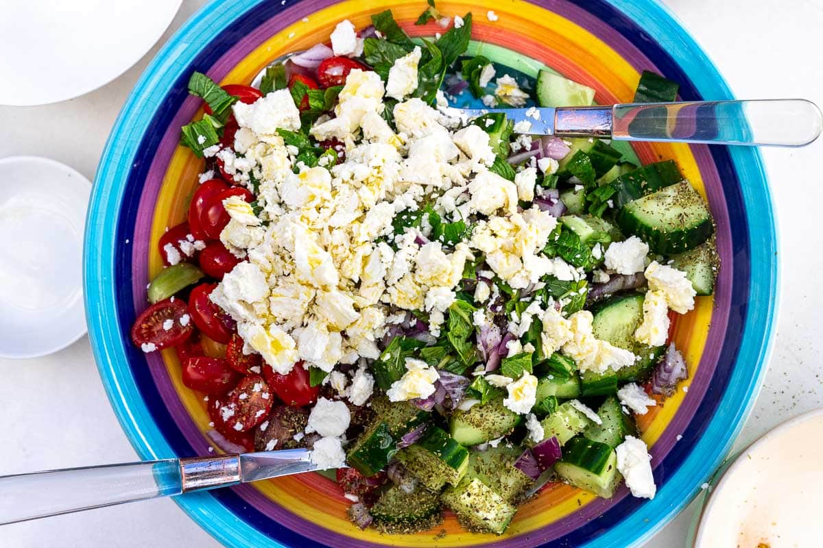 Closeup overhead of a rainbow colored bowl with all the ingredients in it for the salad, with salad servers.