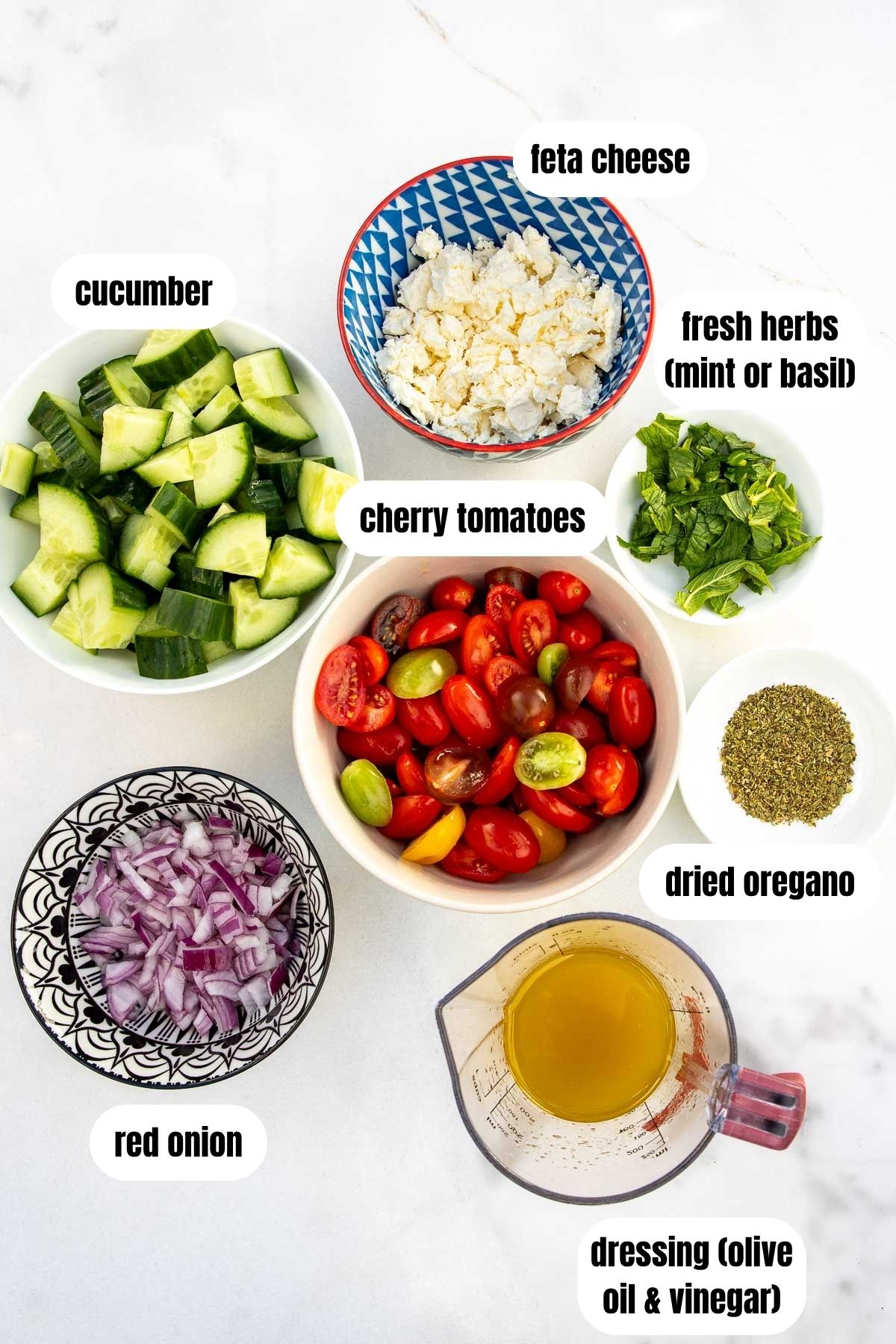 All the ingredients needed to make a cucumber, tomato and feta salad, labelled, including halved cherry tomatoes, cucumber pieces, chopped red onion, crumbled feta, dried oregano, fresh mint, and an olive oil and vinegar dressing.