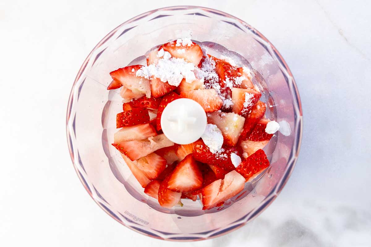 Overhead view of chopped strawberries, powdered sugar and other ingredients in a small food processor.