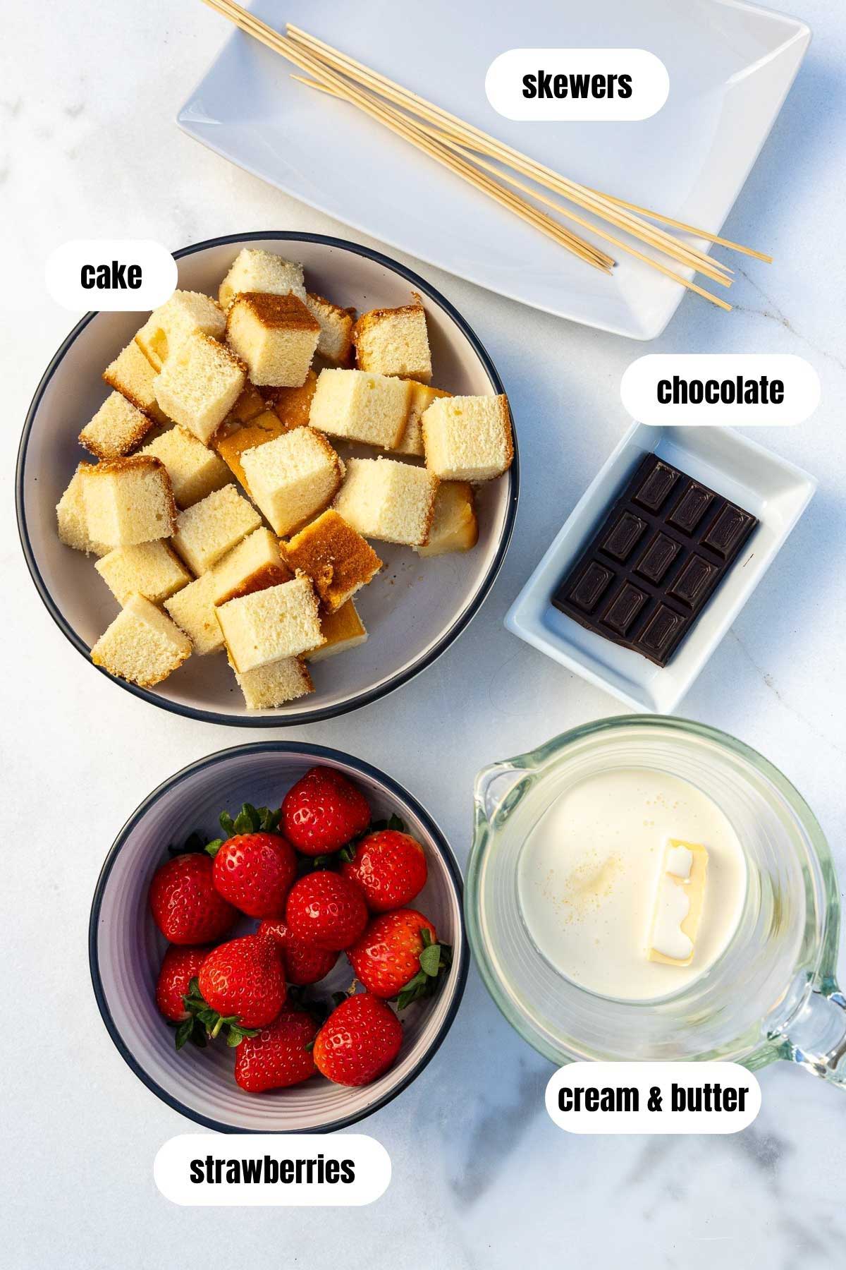 All the ingredients needed to make strawberry and cake skewers, labelled, including wooden skewers, strawberries, cream and butter, cubes of vanilla cake, and chocolate chunks.