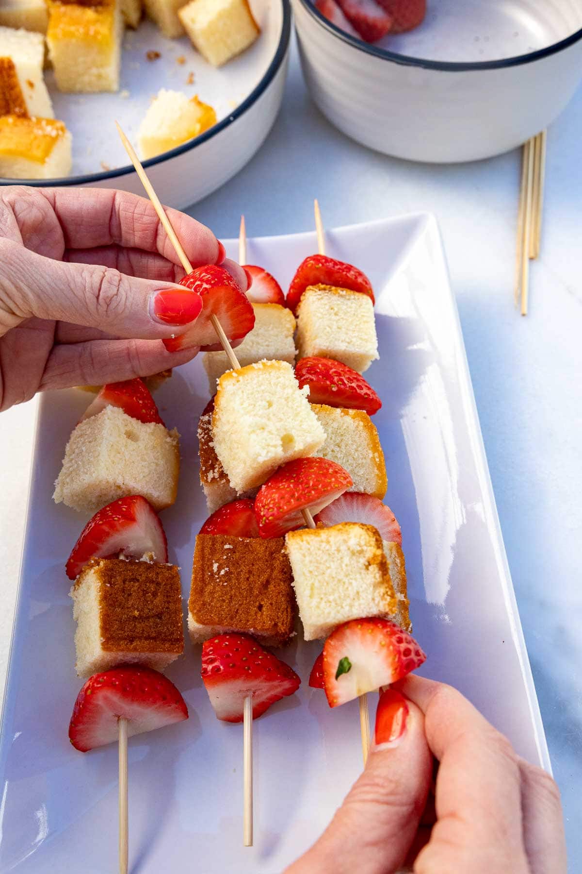 Someone's hands threading half strawberries and cubes of cake onto a skewer, with more strawberry skewers below on a plate.