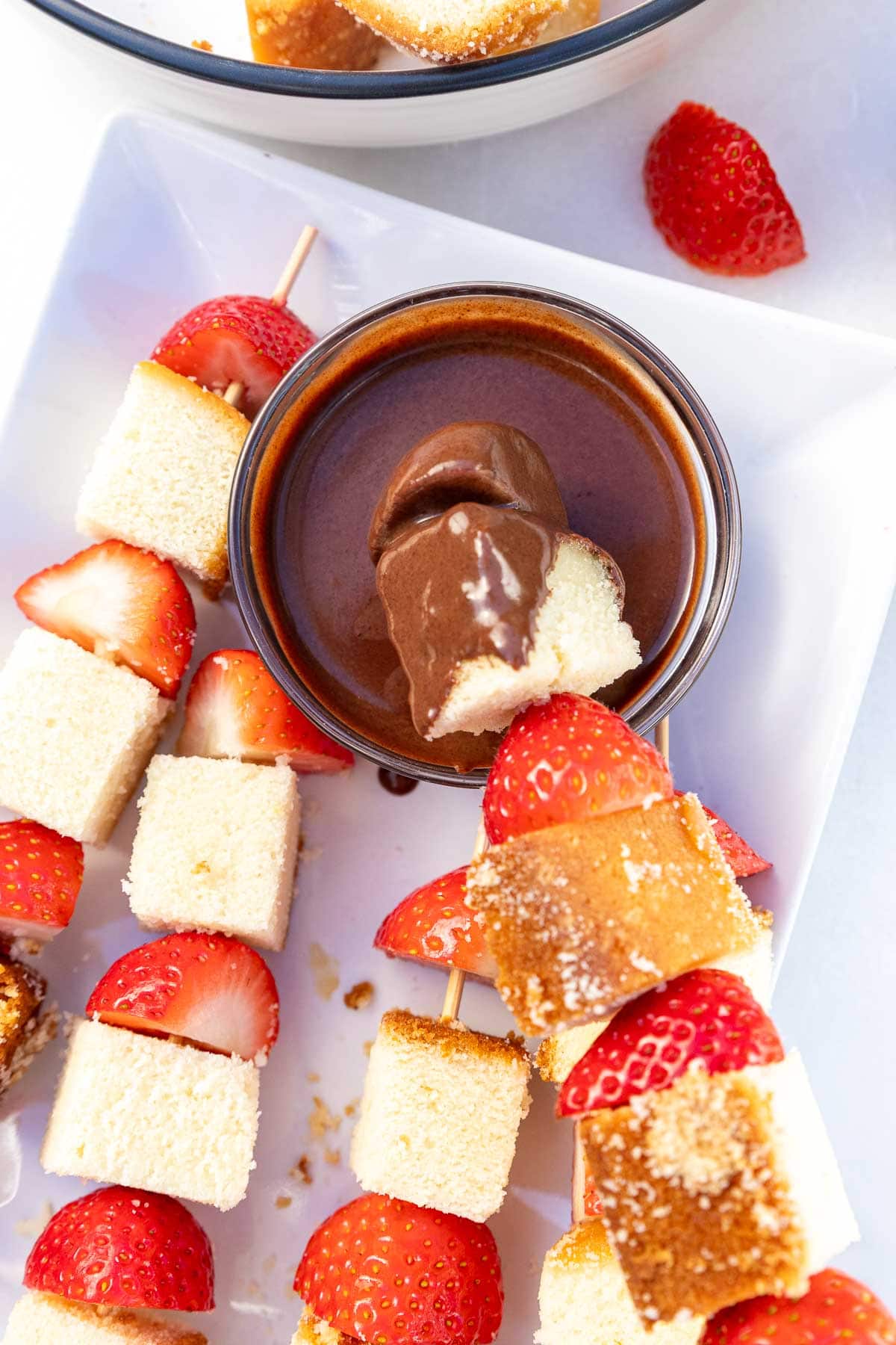 Closeup of half strawberries and cubes of cake threaded onto wooden skewers, one with the end dipped into a small bowl of chocolate sauce.