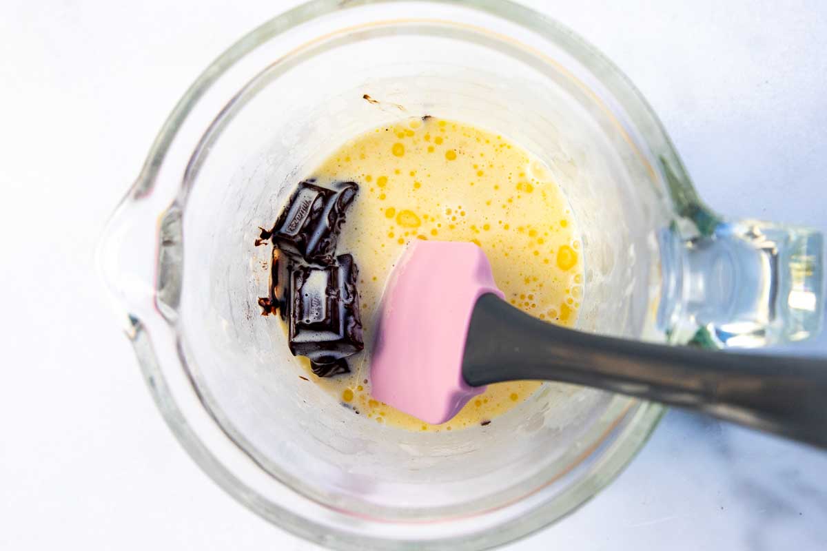 Overhead view of warm cream, butter and chocolate chunks in a glass jug with a pink spatula.