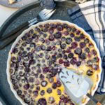 Overhead view of a blueberry clafoutis in a round white dish with a slice cut out of it and a cake slice.
