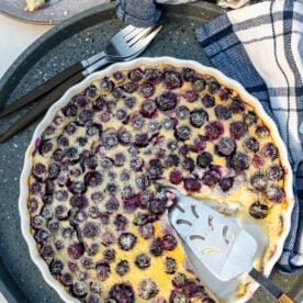 Overhead view of a blueberry clafoutis in a round white dish with a slice cut out of it and a cake slice.