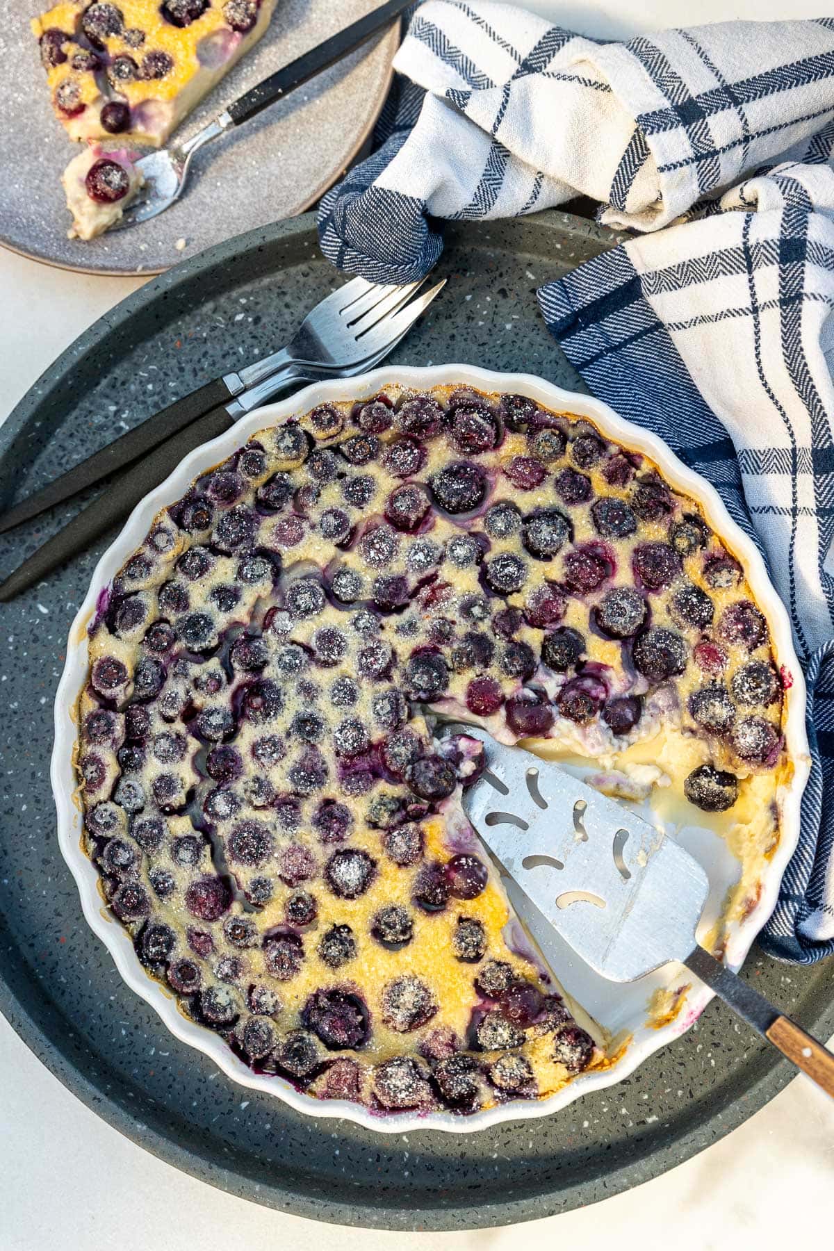 Overhead view of a blueberry clafoutis in a round white dish with a slice cut out of it and a cake slice.