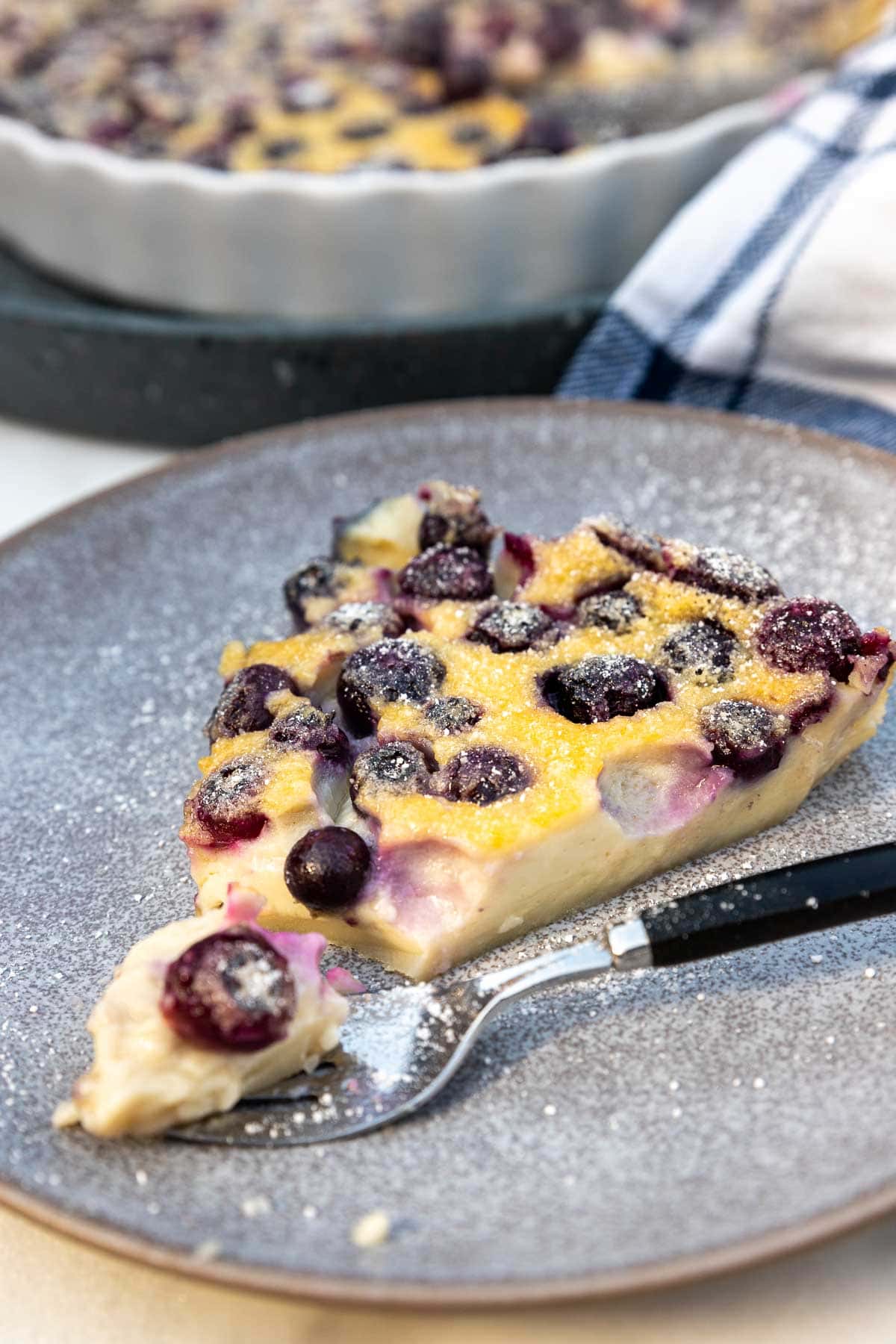 A closeup of a slice of the clafoutis on a medium blue speckled plate with a black-handled fork taking the first bite.