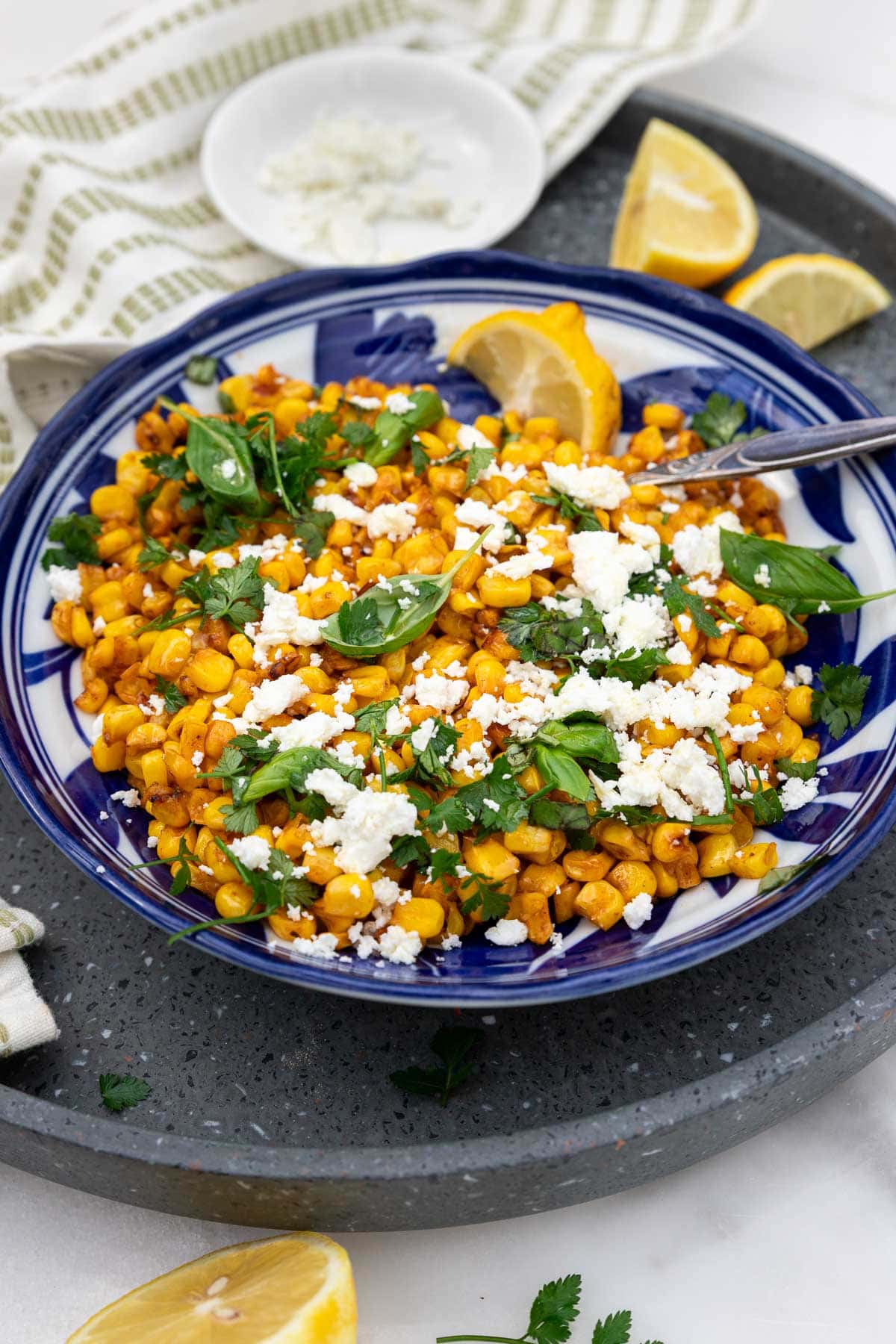 A blue dish with sauteed corn and feta and fresh herbs on a grey tray with a green and cream tea towel at the side.