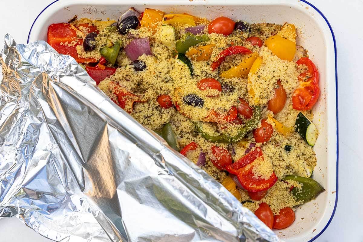 Overhead view of roasted vegetables with raw couscous and broth poured over and aluminium foil tucked over the pan ready to bake.