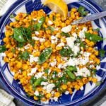 An overhead view of a blue dish with sauteed corn, feta cheese and herbs, on a grey tray and with a cream and green tea towel.