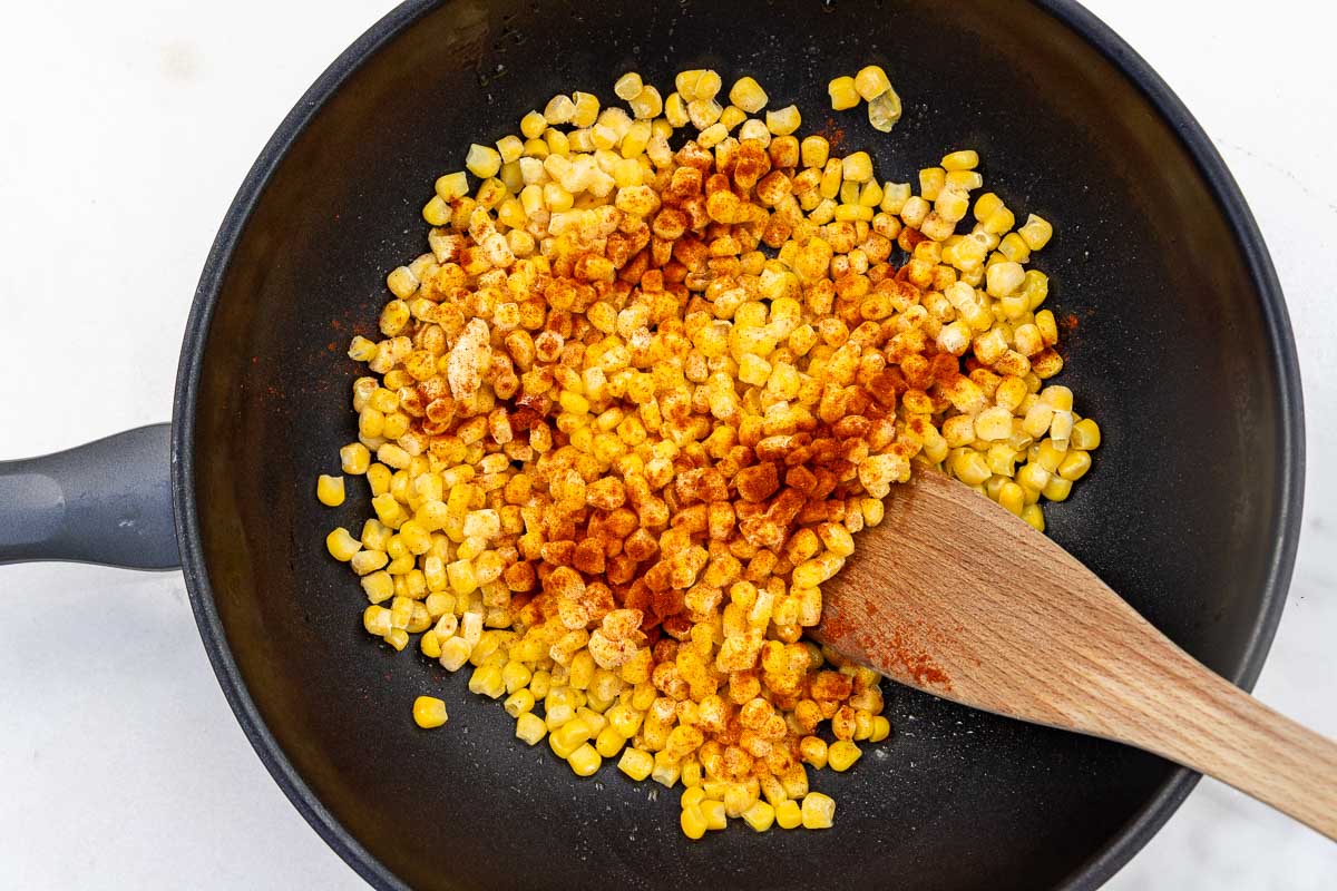 Overhead view of frozen corn kernels in a pan with paprika sprinkled over and a wooden spoon.