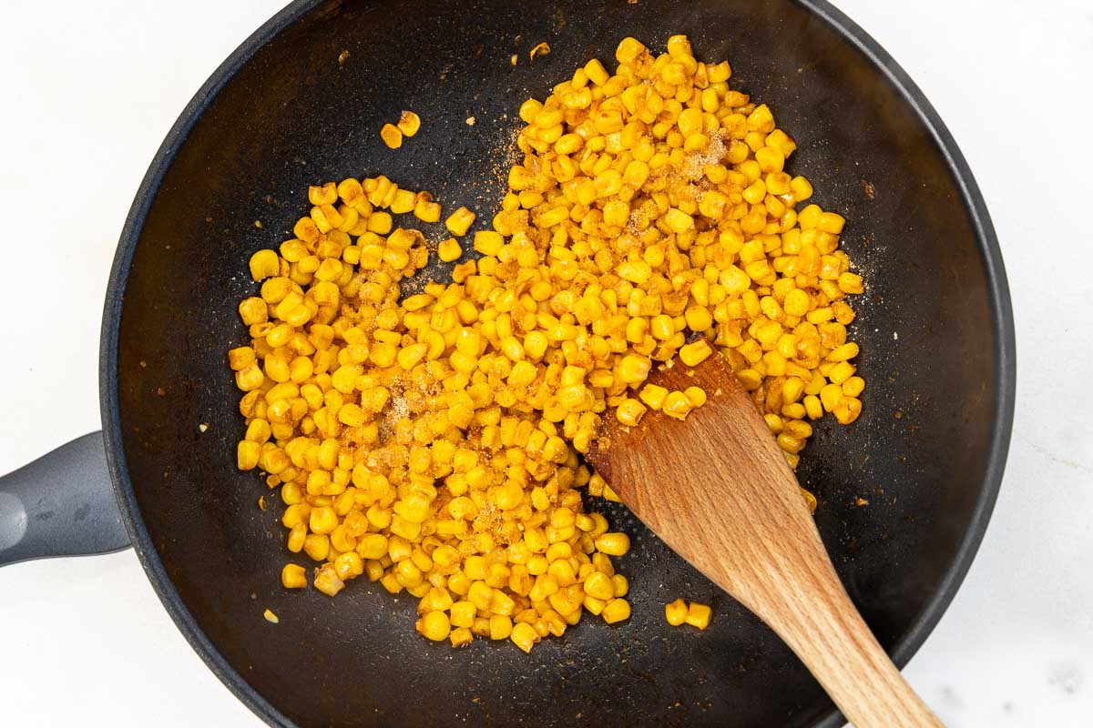 Overhead view of frozen corn kernels in a wok style pan with seasoning sprinkled over and a wooden spoon.